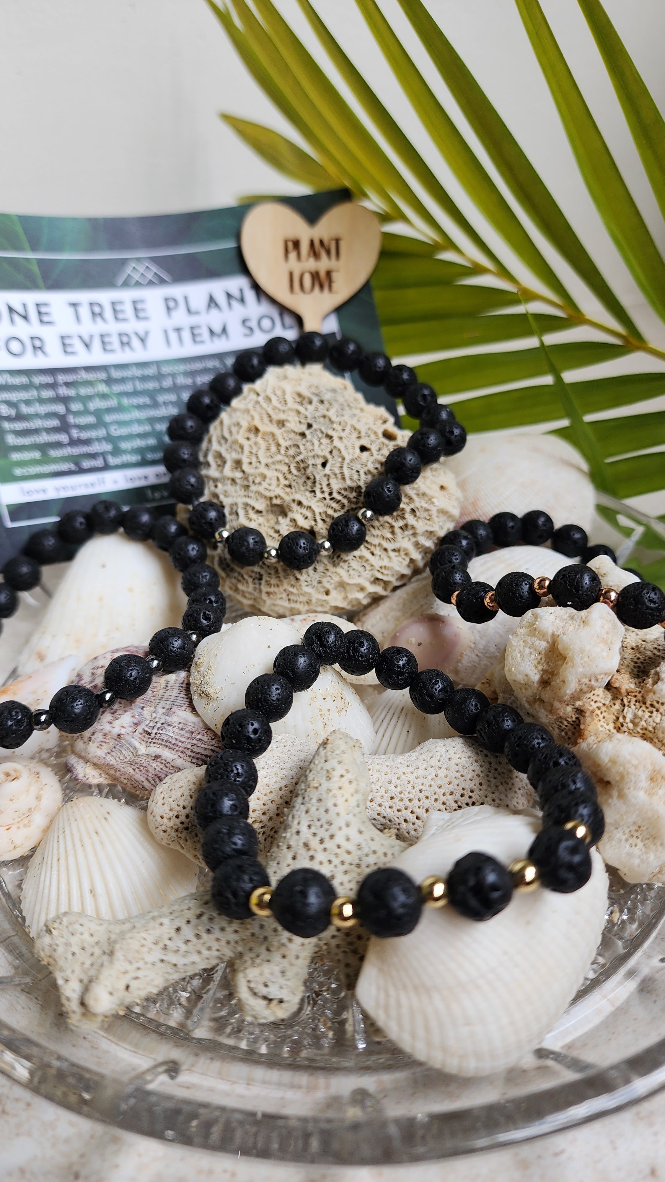 Four lava stone bracelets resting along shells and beach rocks, with a palm leaf and 'lovelevel' card in the background.