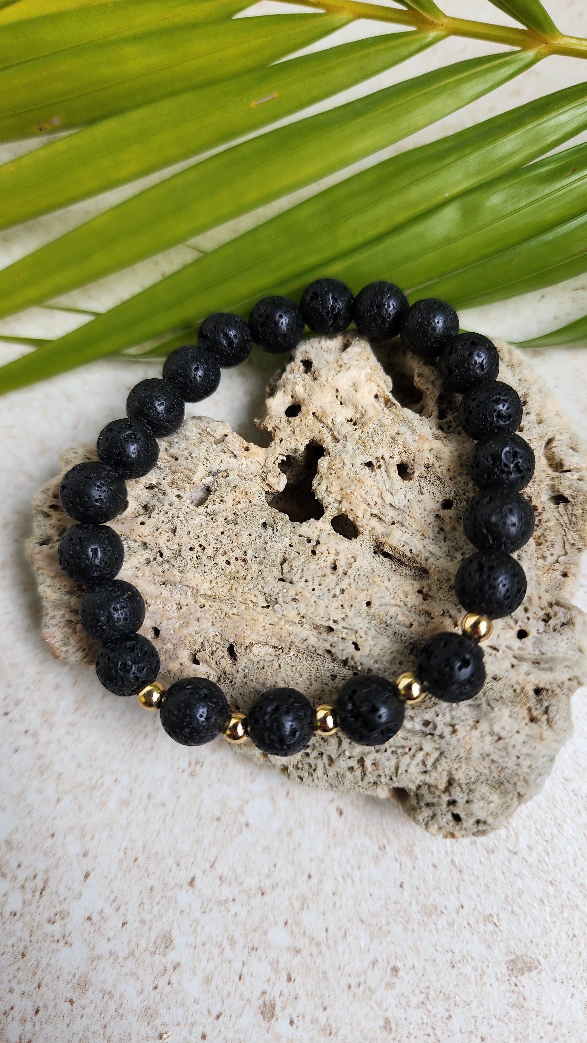 A lava stone bracelet with 5 golden beads resting along a heart shaped stone with a leaf in the background.