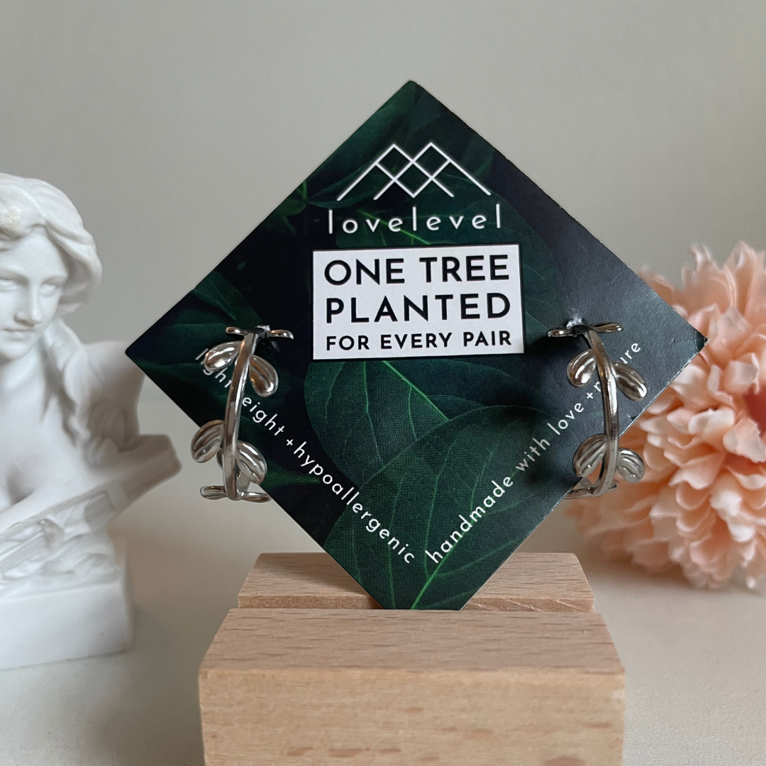 Silver hoop earrings with a vine design on a card with 'lovelevel' branding. In the background rests a statue and pink flower.