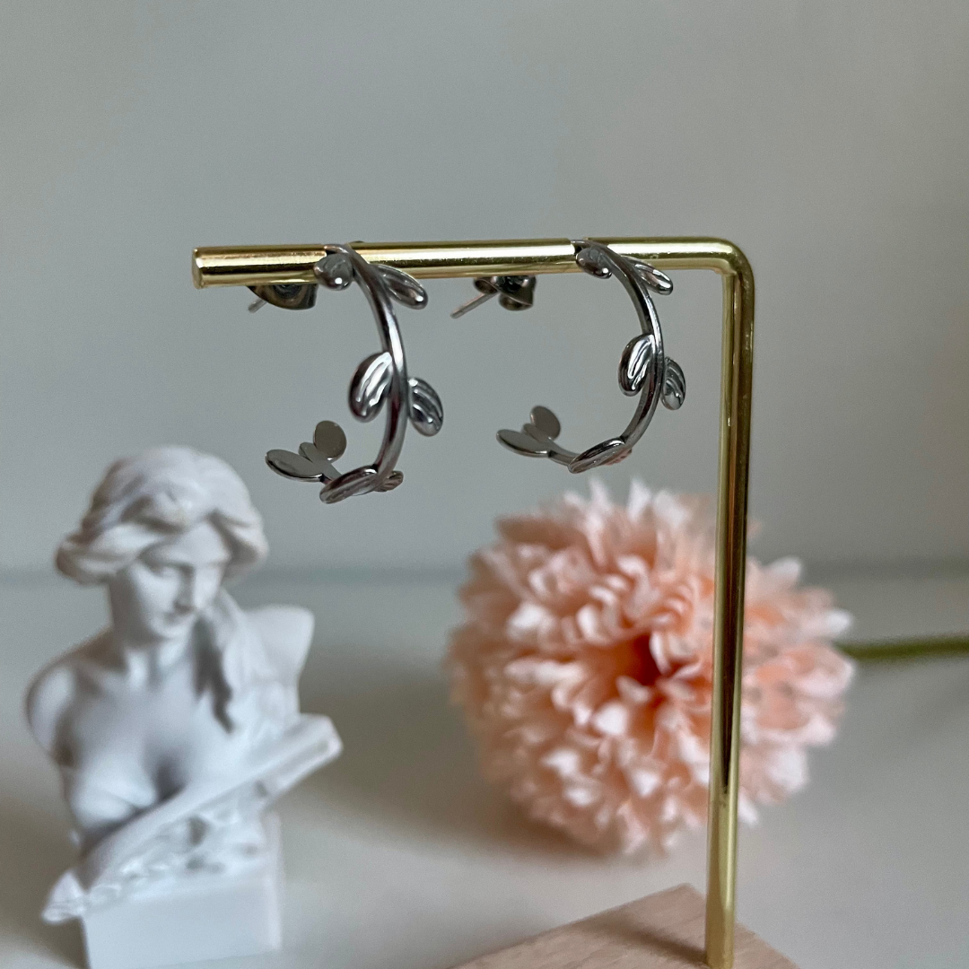 Silver hoop earrings with a vine design hanging from a golden bar with a statue and pink flower in the background.