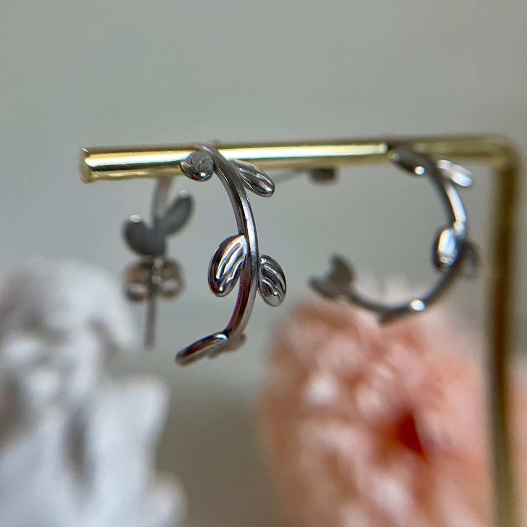Silver hoop earrings with a vine design draped from a gold bar against a blurred background.