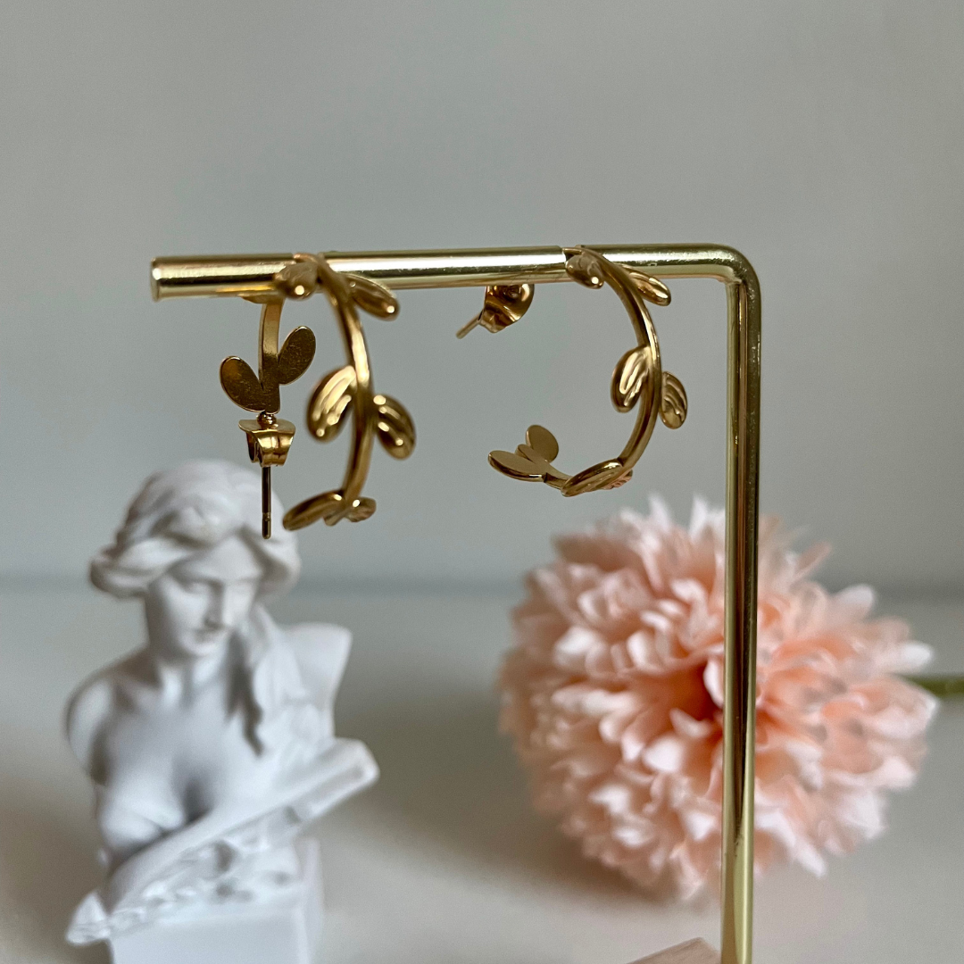 Golden hoop earrings with a vine design draped from a gold bar. A statue and pink flower sit in the background.