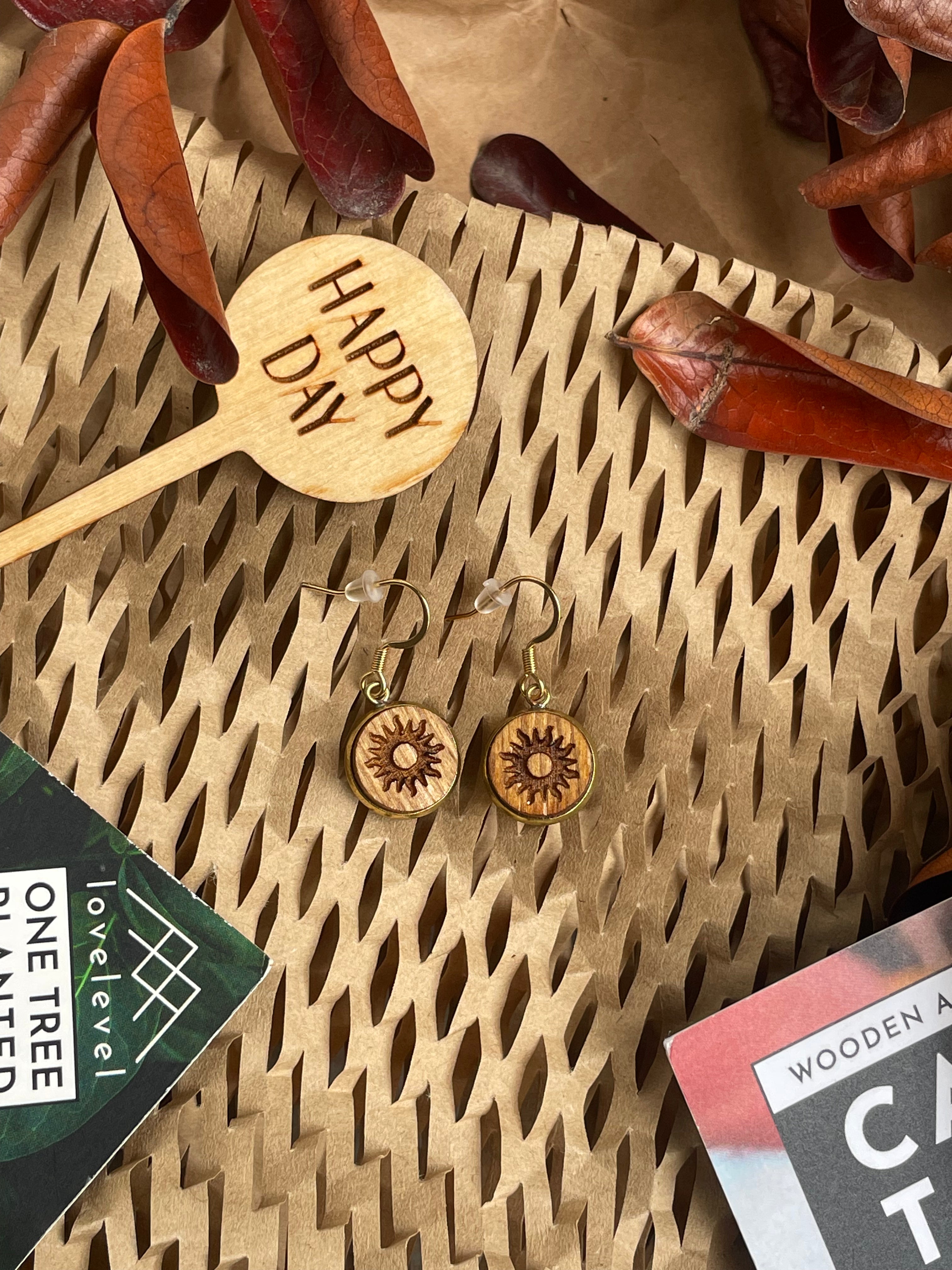Wooden dangle earrings with a sun design against a natural background with a 'Happy Day' plant marker.