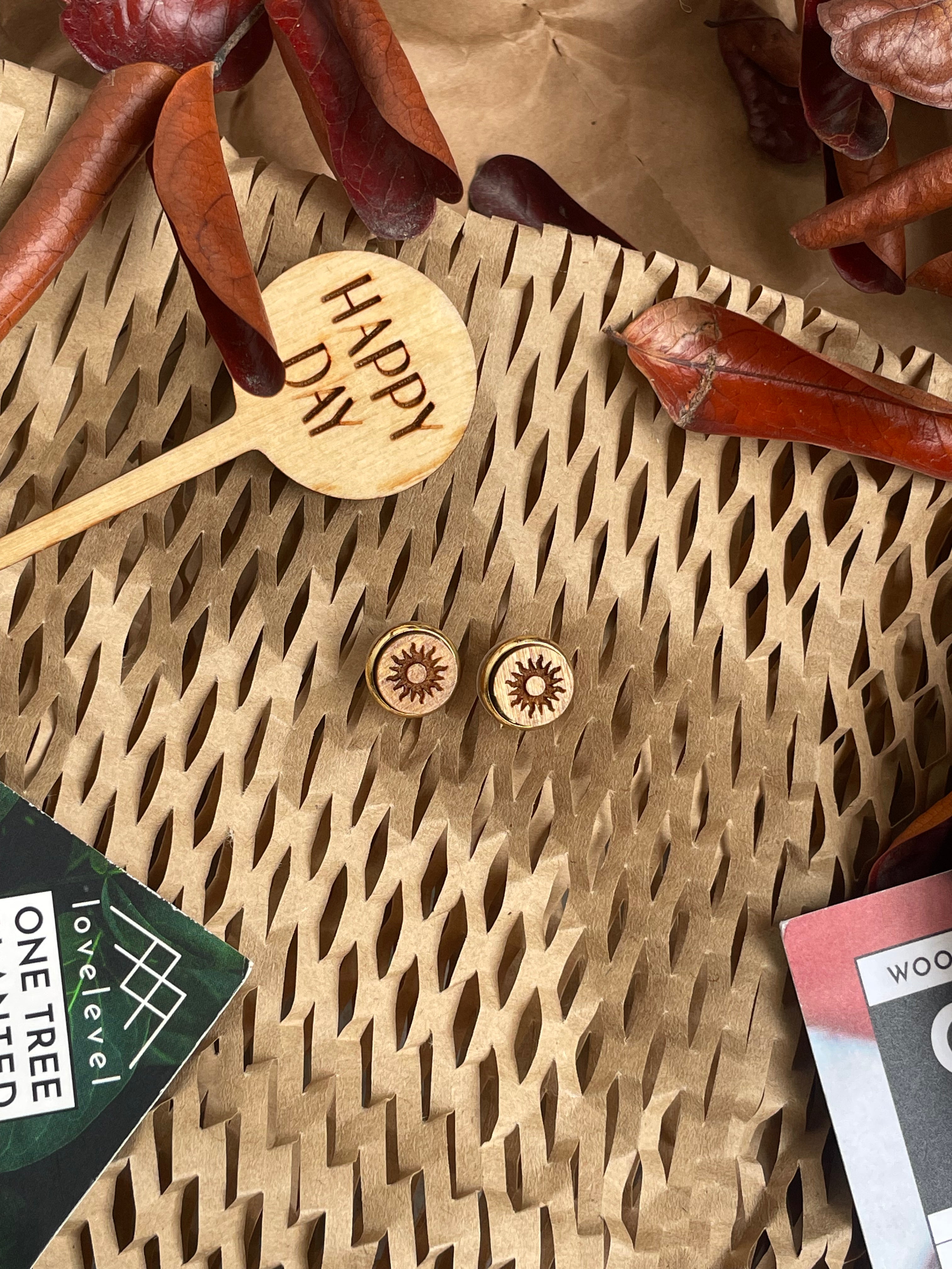 Wooden stud earrings with a sun design against a natural background with a "Happy Day" plant marker.