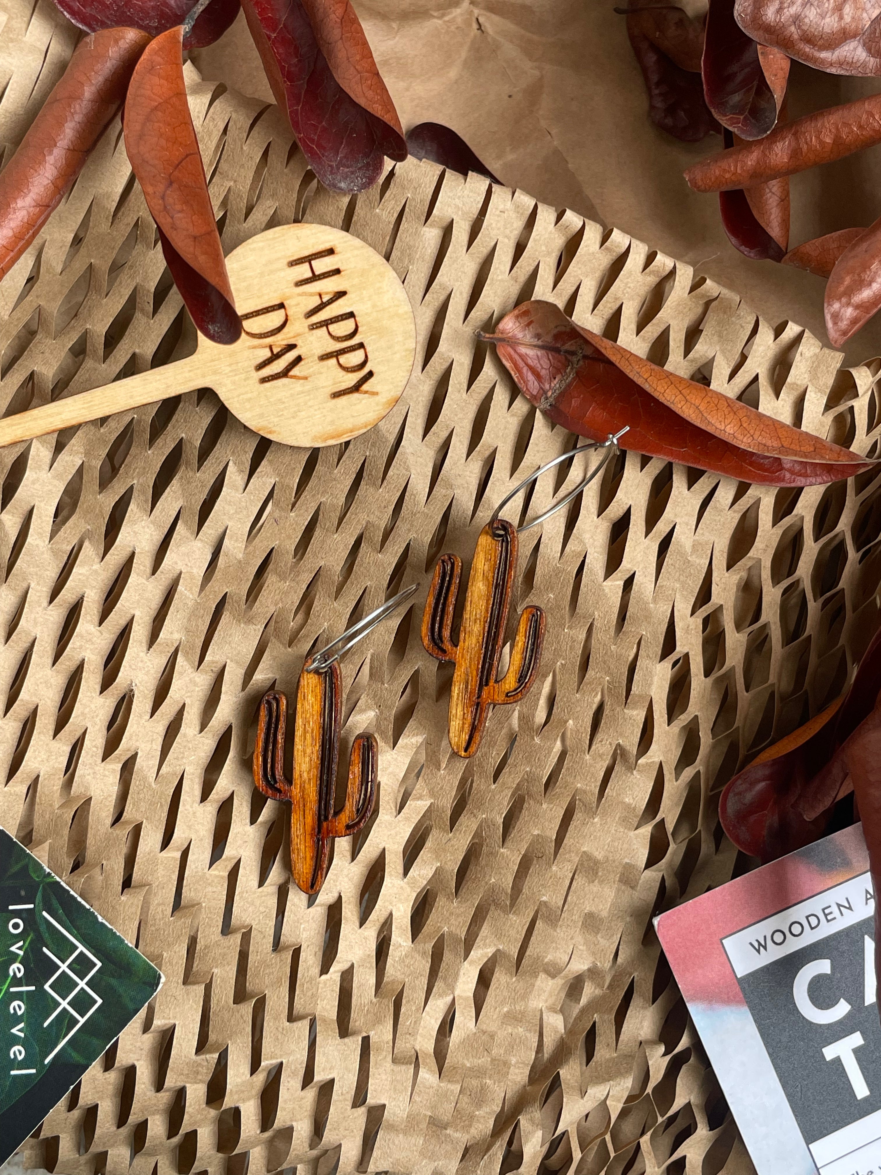 Wooden cactus earrings on a natural background with dried leaves and a "Happy Day" plant marker.