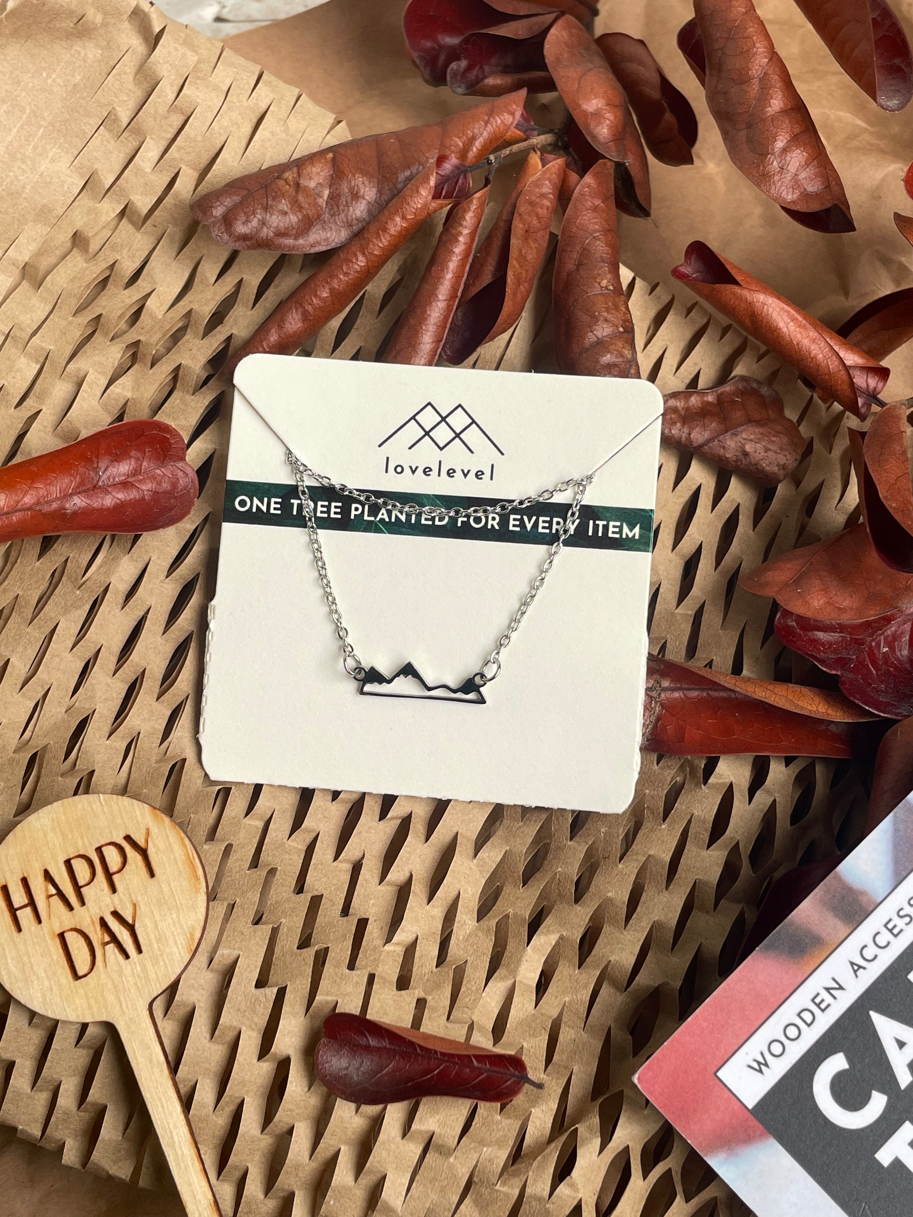 Metal mountain peak necklace on a card with 'lovelevel' branding against a natural background with dried leaves and a "Happy Day" plant marker.