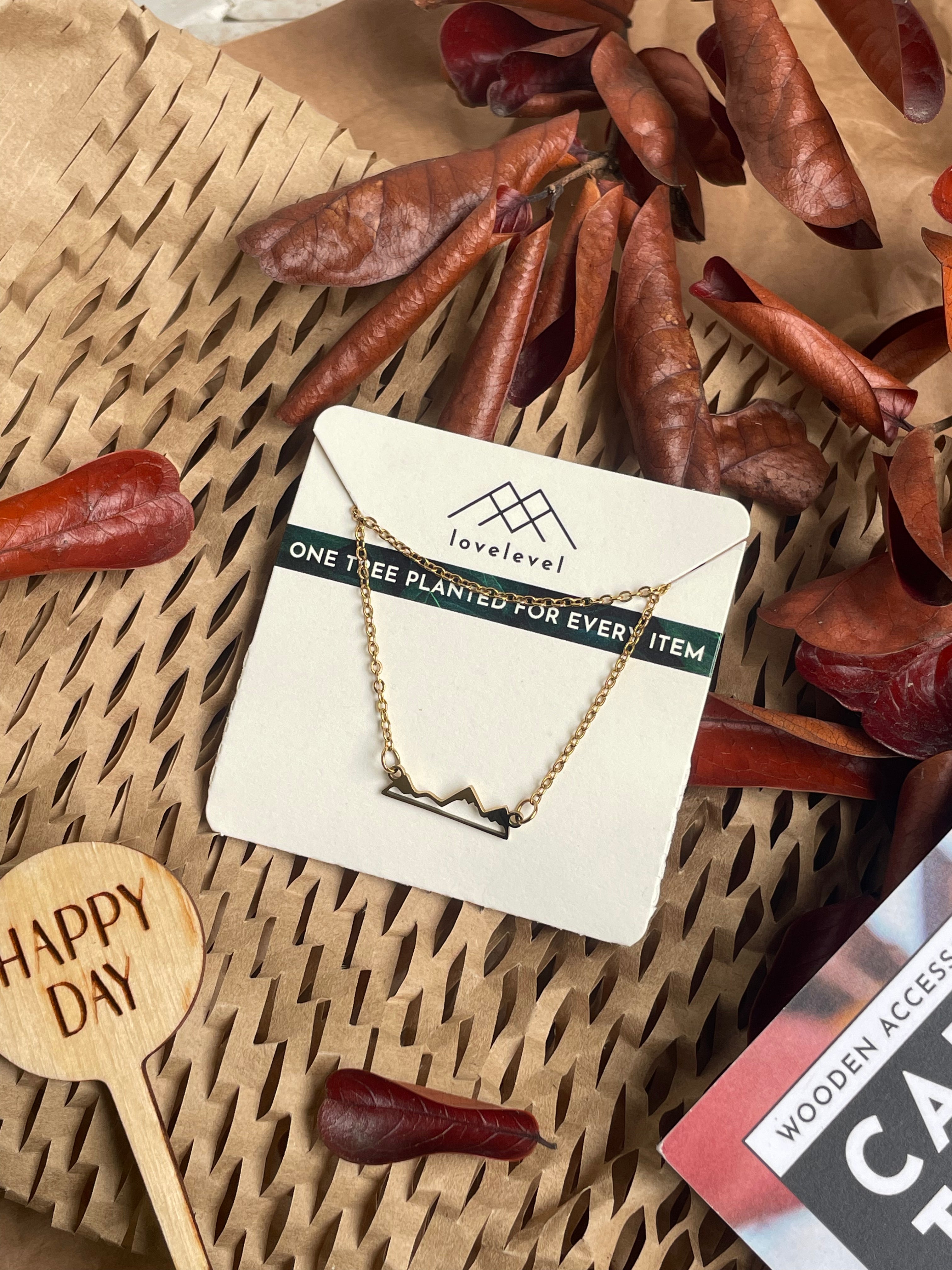 Metal mountain peak necklace on a card with 'lovelevel' branding against a natural background with dried leaves and a "Happy Day" plant marker.