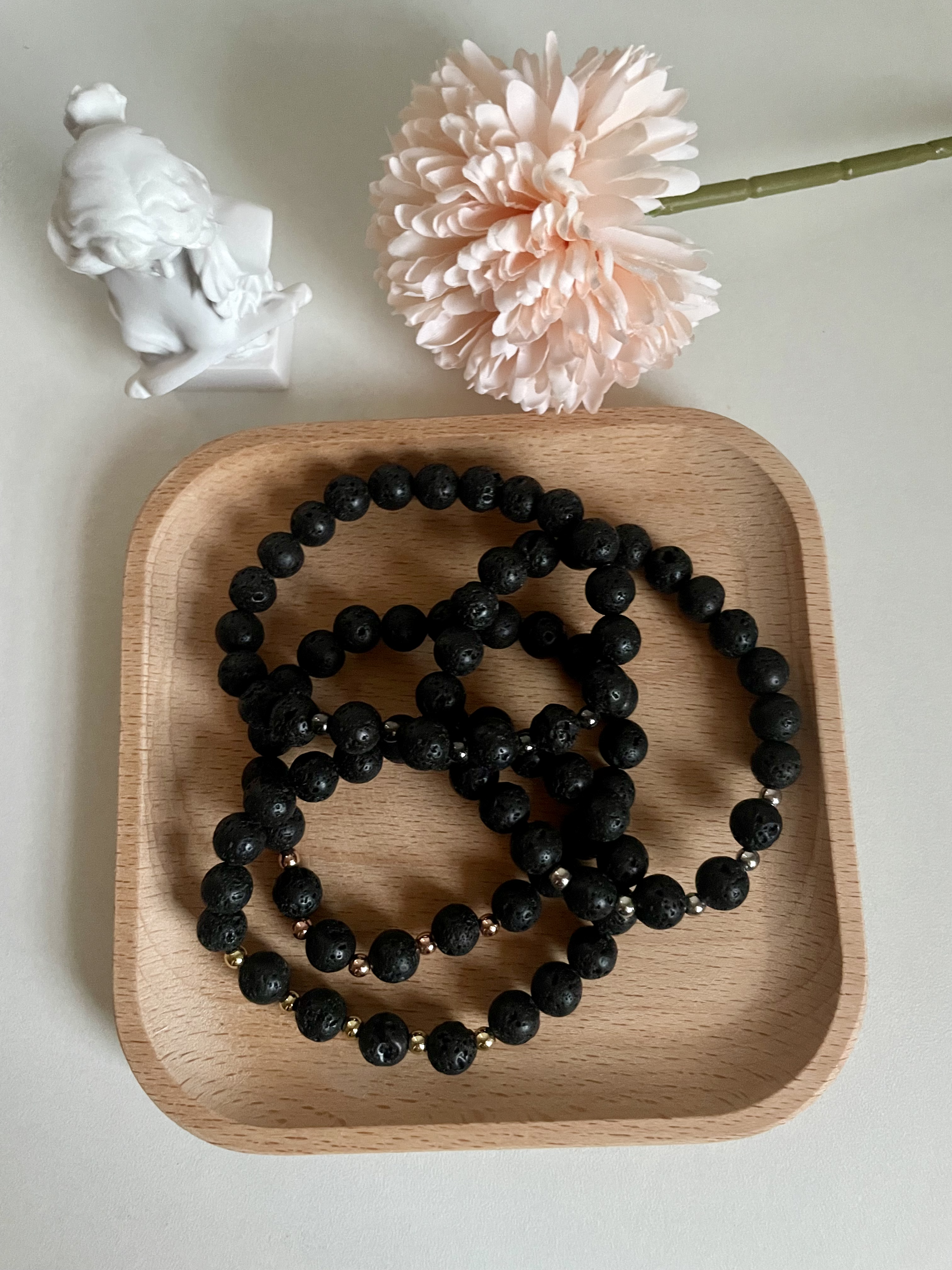 Four lava stone bracelets on a wooden tray in front of a statue and pink flower with the stem.