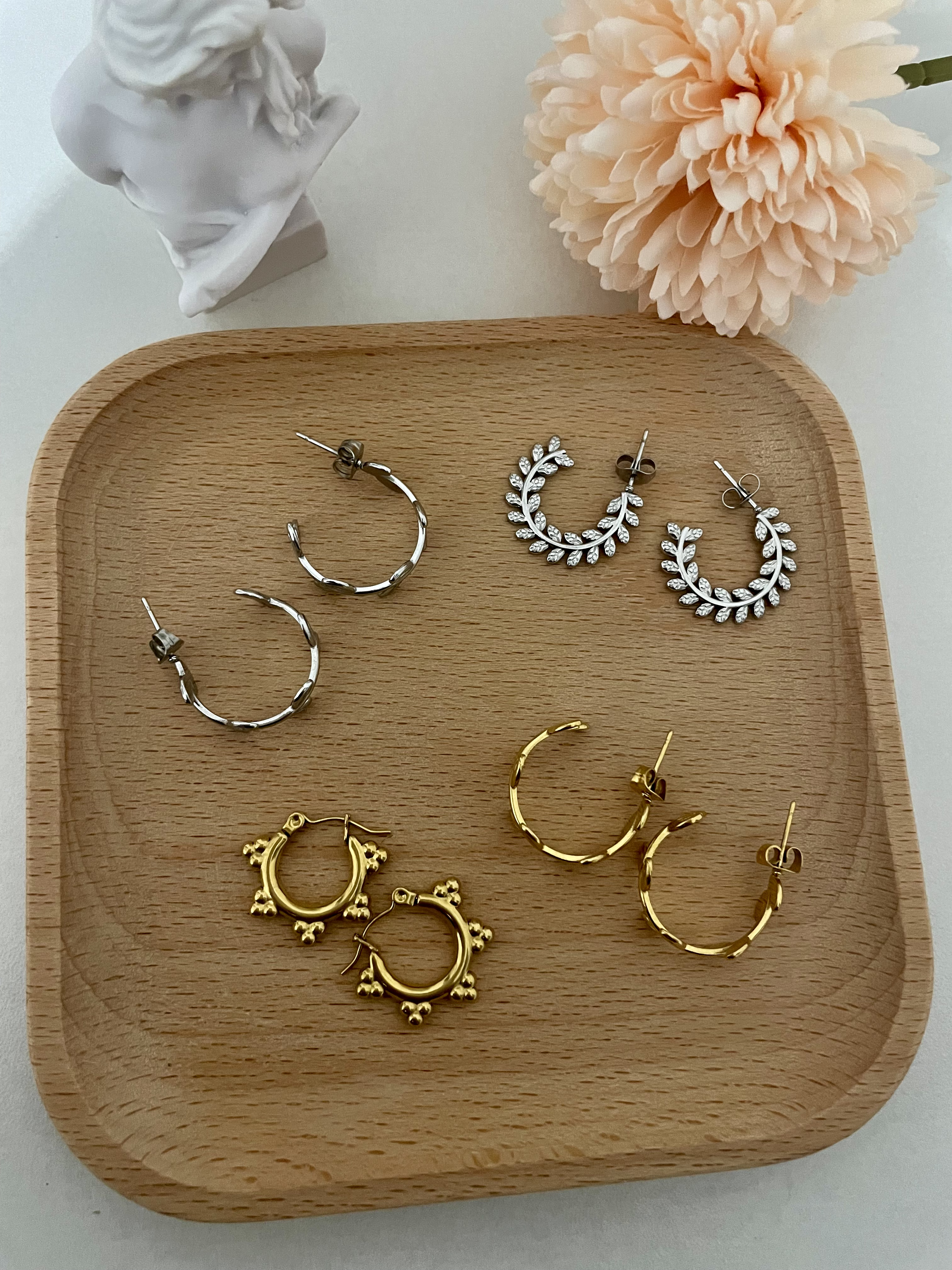 Four pairs of hoop earrings (two silver and two gold) on a wooden tray in front of a statue and pink flower.