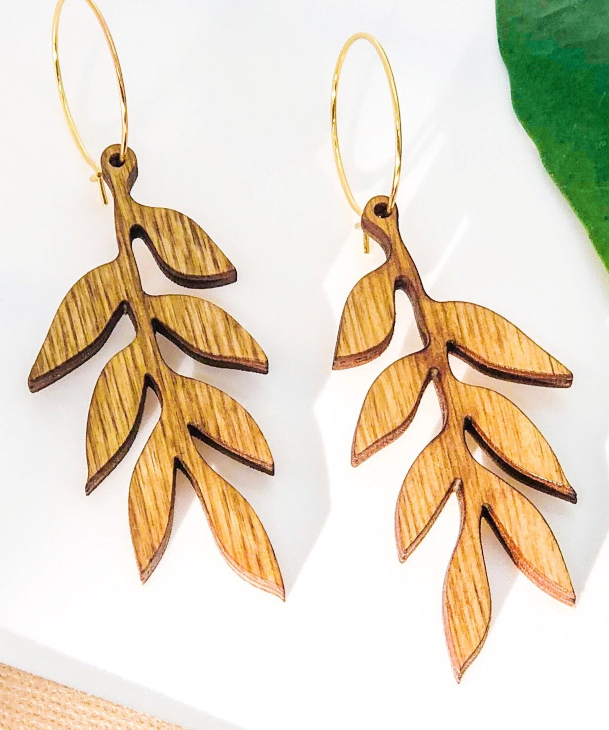 Wooden leaf-shaped earrings on a white background with a green leaf and beige fabric.