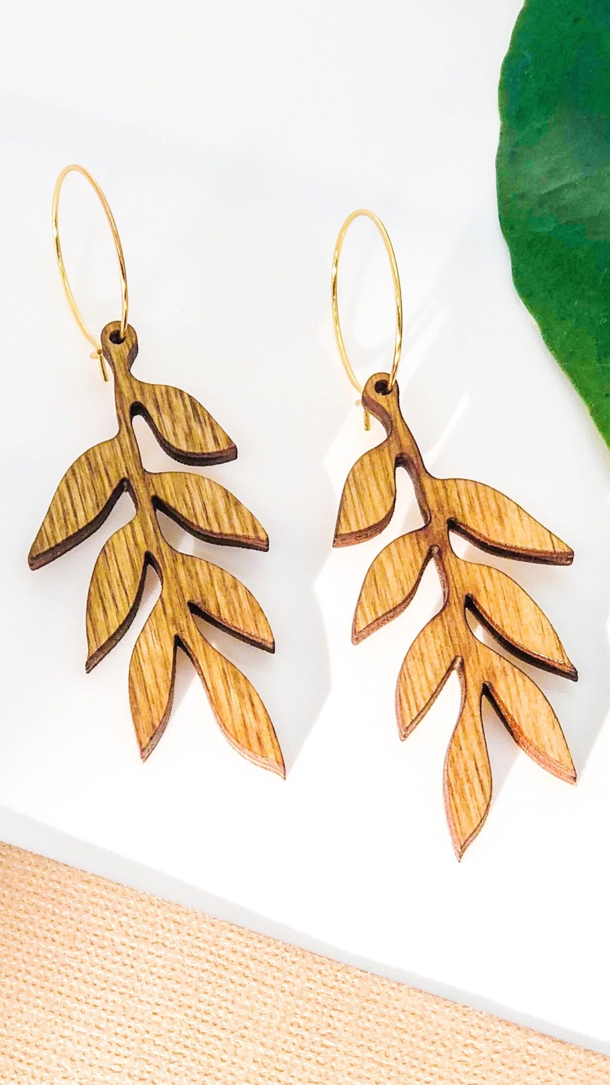 Wooden leaf-shaped earrings on a white background with a green leaf and beige fabric.