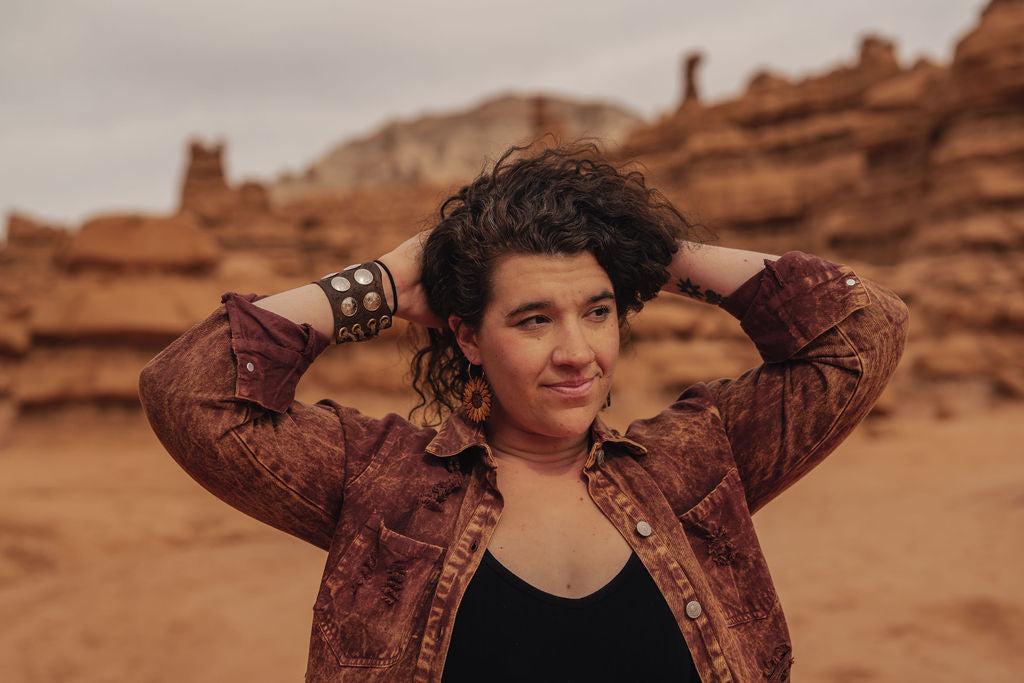 Person with curly hair and sunflower earrings standing in a desert landscape with rock formations.
