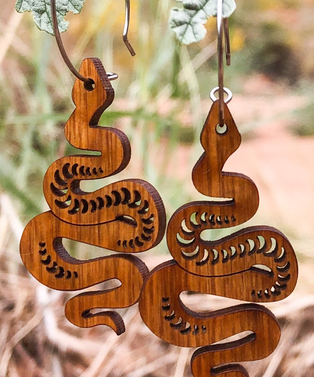 Wooden snake-shaped earrings hanging on a branch with dried leaves and blurred natural background