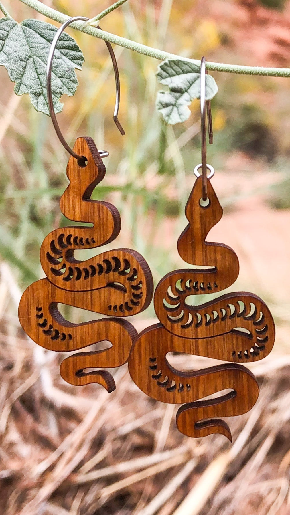 Wooden snake-shaped earrings hanging on a branch with dried leaves and blurred natural background