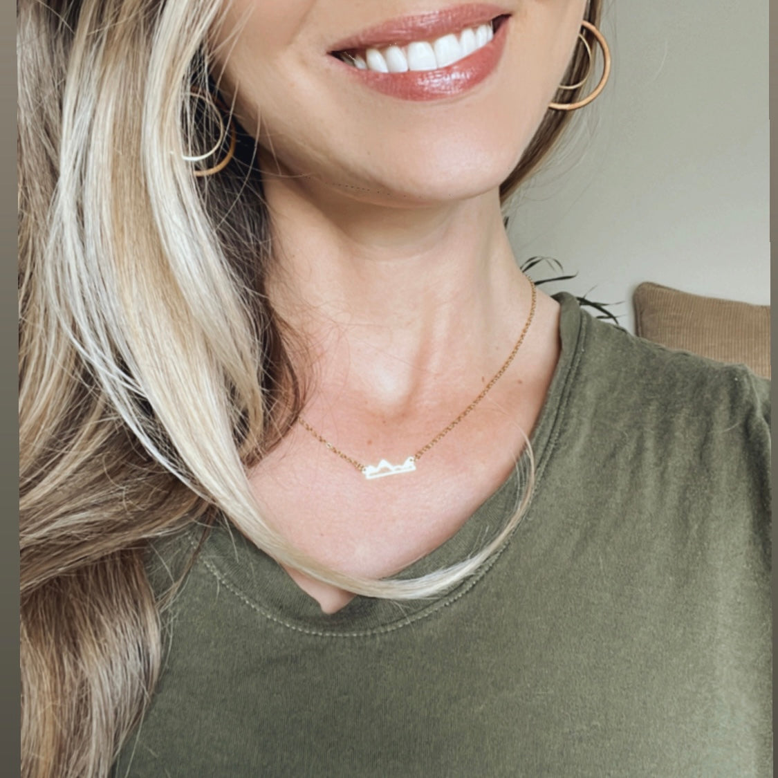 Smiling woman wearing large wooden hoop earrings with metal rings in the hoop, and a golden necklace with a small mountain silhouette charm.