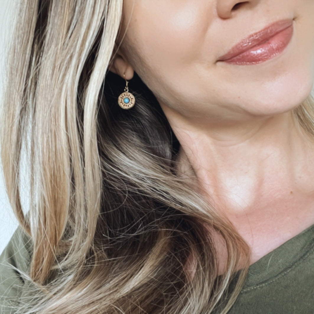 Smiling blond woman wearing a wooden dangle earring with a bright blue stone in the center of the earring.