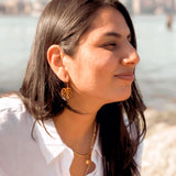 Smiling woman wearing wooden leaf earrings on a beach.