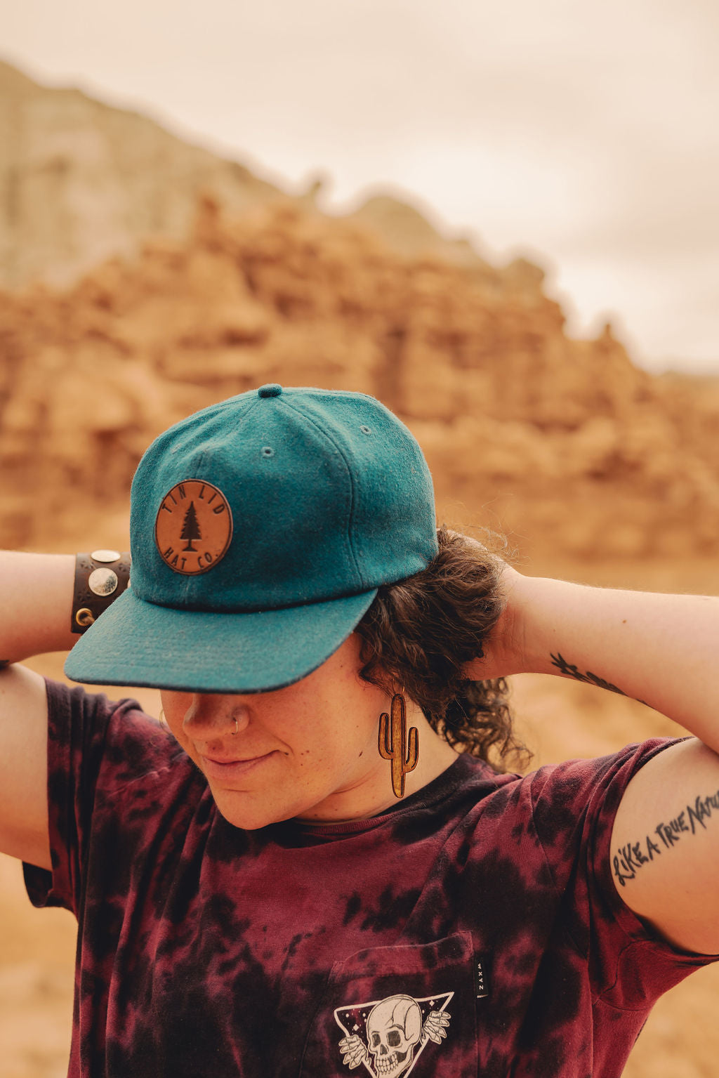 Person in the desert wearing a blue cap and wooden cactus earrrings.