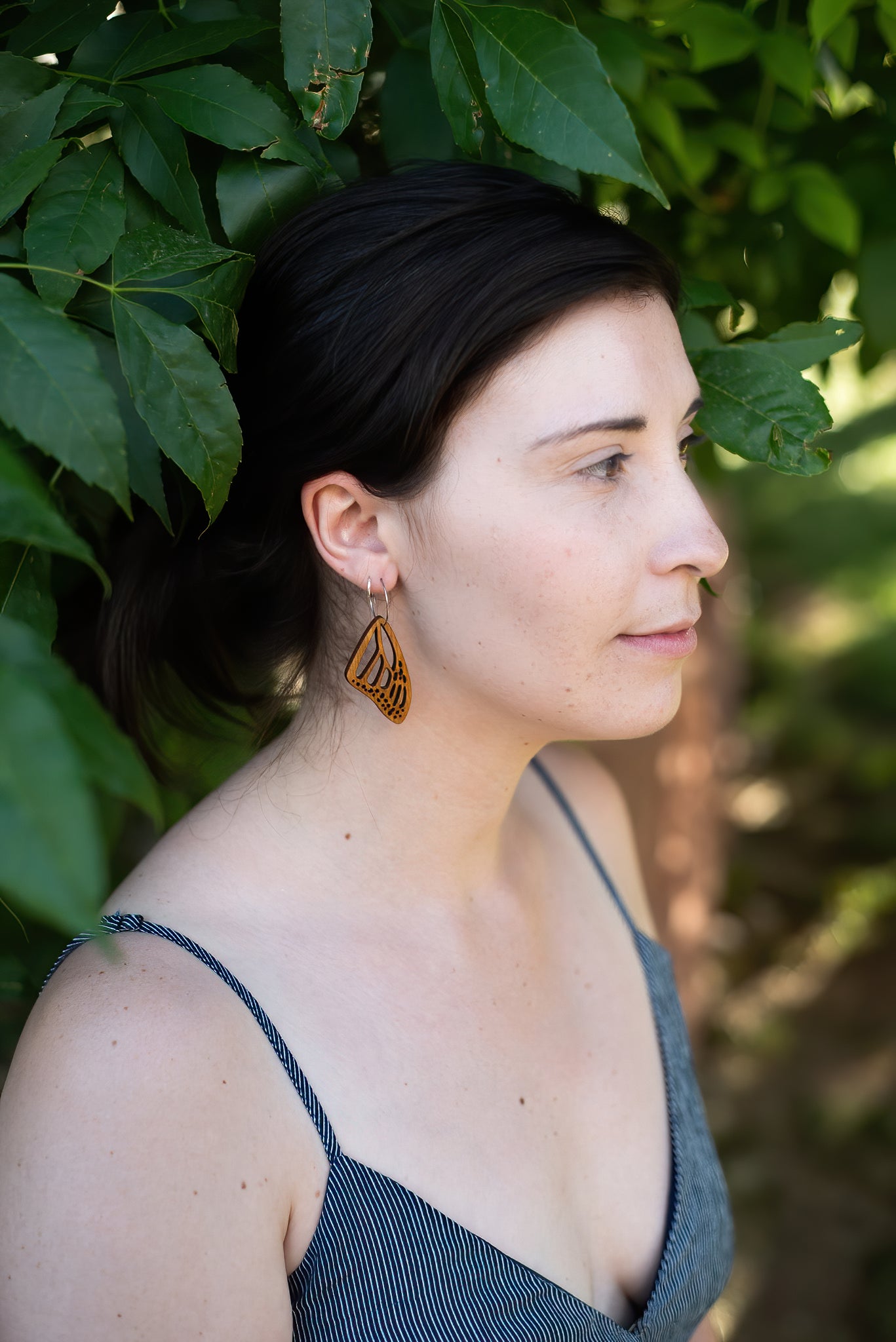 Woman wearing wooden butterfly earring looking off to the side with leaves in the background.