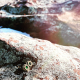Pair of wooden stud earrings with a green stone in the center sitting along beach rocks.
