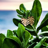 A pair of wooden leave earrings sitting on a leaf in front of an ocean sunset.