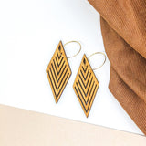 Wooden diamond earrings with geometric shapes carved into them in front of brown fabric.