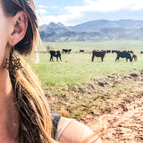 Close up of woman's ear wearing wooden lotus earring in front of a mountain ranch.
