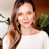 Smiling woman wearing wooden crescent earrings in front of plants and a door.