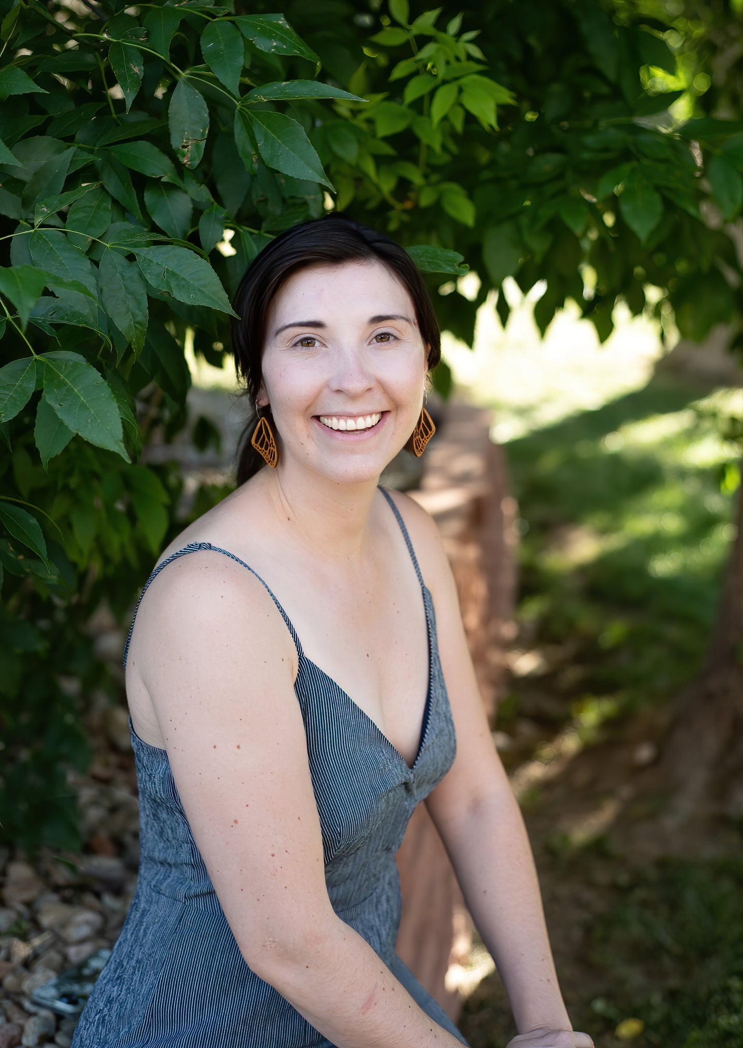Smiling woman sitting under a tree wearing wooden earrings in the shape of butterfly wings.