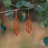 Pair of wooden earrings carved into a long diamond shape hanging from a stick in the woods.