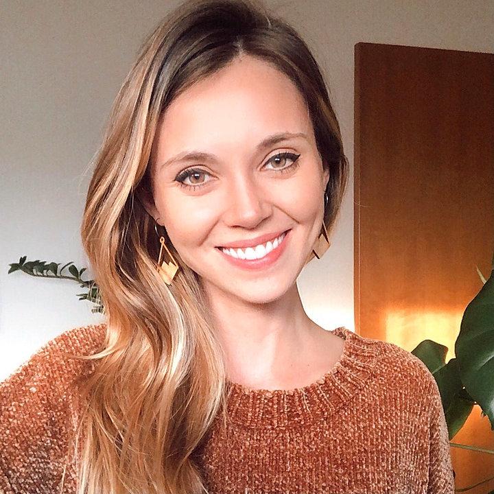 Smiling woman wearing wooden diamond earrings with mountain peaks carved in, wearing an orange sweater in front of plants.