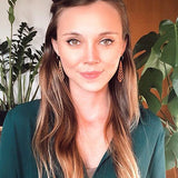 Smiling woman wearing wooden slim diamond earrings wearing a green sweater in front of plants.