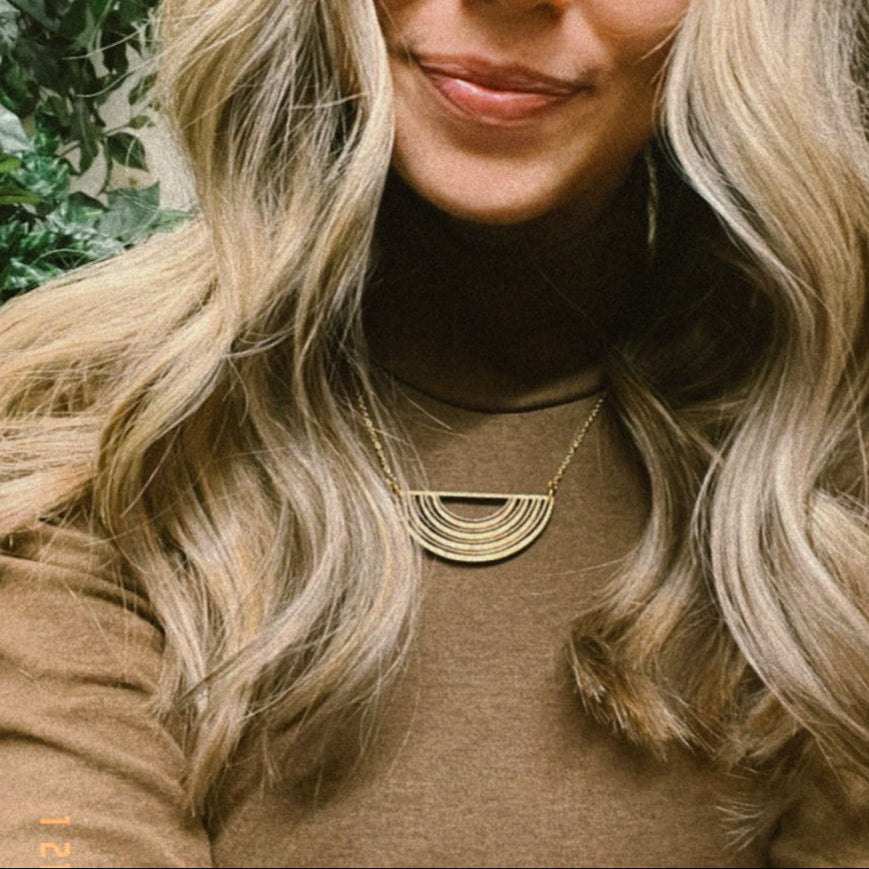 Woman with long blonde hair wearing a brown top and gold necklace with a half circle wooden pendant, standing outdoors with greenery in the background.