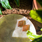 Pair of wooden geometric diamond earrings sitting along a stone plate under leaves.