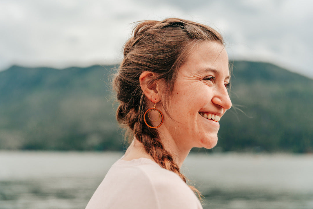 Smiling woman wearing large wooden hoop earring, with a golden ring hanging in the hoop, against a mountain background.