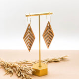 Geometric wooden diamond earrings hanging from a gold bar over wheat plants.