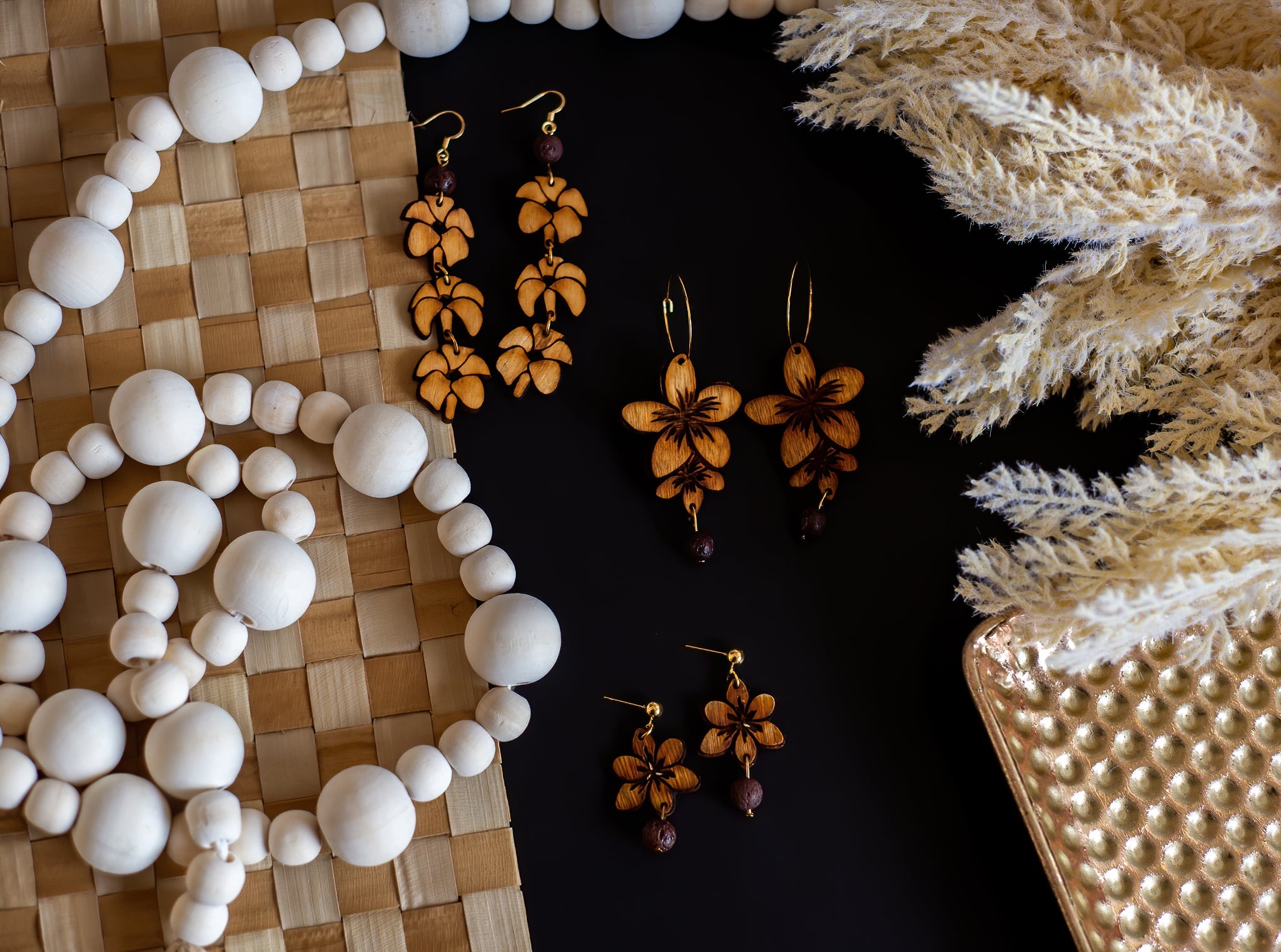 Colletion of wooden flower earrings with lava stone beads on a black table with white and beige table cloths.