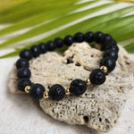 Lava stone bracelet resting along a heart shaped stone with a palm leaf in the background.