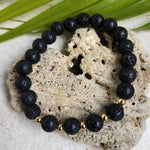 A lava stone bracelet with 5 golden beads resting along a heart shaped stone with a leaf in the background.