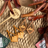 Mountain earrings against a natural background with dried leaves and a "Happy Day" plant marker.