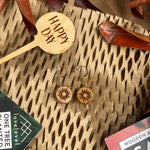 Wooden dangle earrings with a sun design against a natural background with a 'Happy Day' plant marker.