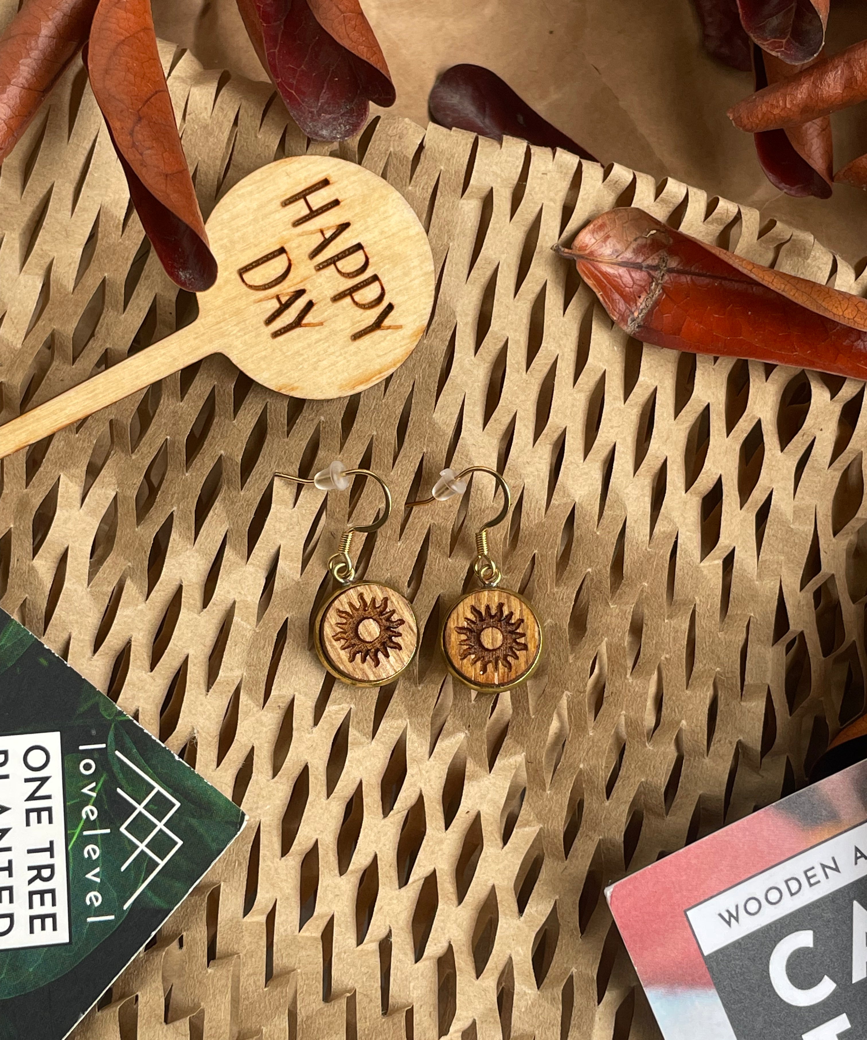 Wooden dangle earrings with a sun design against a natural background with a 'Happy Day' plant marker.