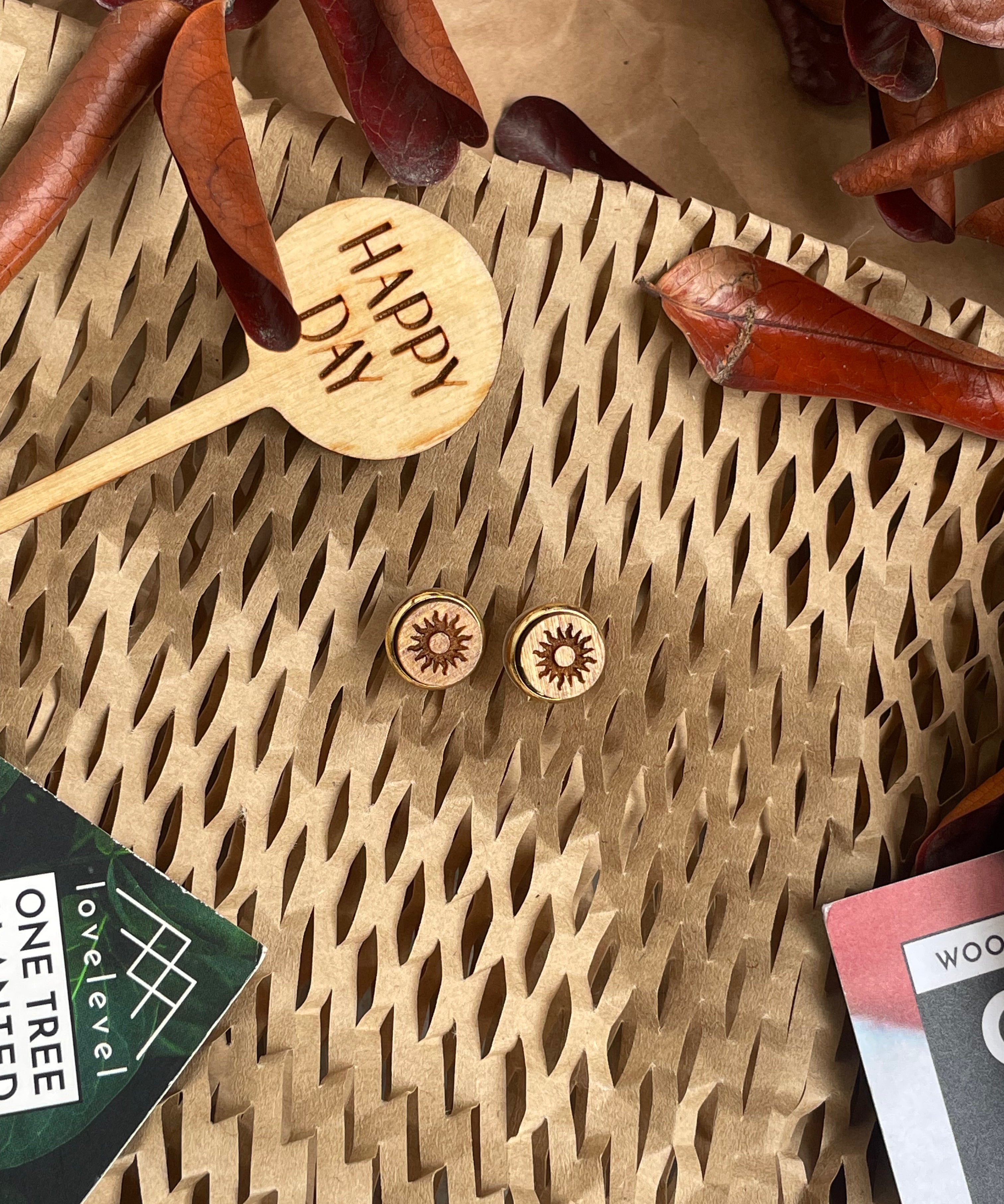 Wooden stud earrings with a sun design against a natural background with a "Happy Day" plant marker.