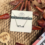 Metal mountain peak necklace on a card with 'lovelevel' branding against a natural background with dried leaves and a "Happy Day" plant marker.