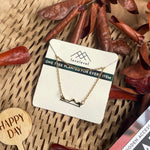 Metal mountain peak necklace on a card with 'lovelevel' branding against a natural background with dried leaves and a "Happy Day" plant marker.