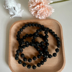 Four lava stone bracelets on a wooden tray in front of a statue and pink flower with the stem.