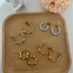 Four pairs of hoop earrings (two silver and two gold) on a wooden tray in front of a statue and pink flower.