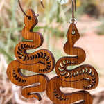 Wooden snake-shaped earrings hanging on a branch with dried leaves and blurred natural background