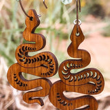 Wooden snake-shaped earrings hanging on a branch with dried leaves and blurred natural background