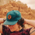 Person in the desert wearing a blue cap and wooden cactus earrrings.